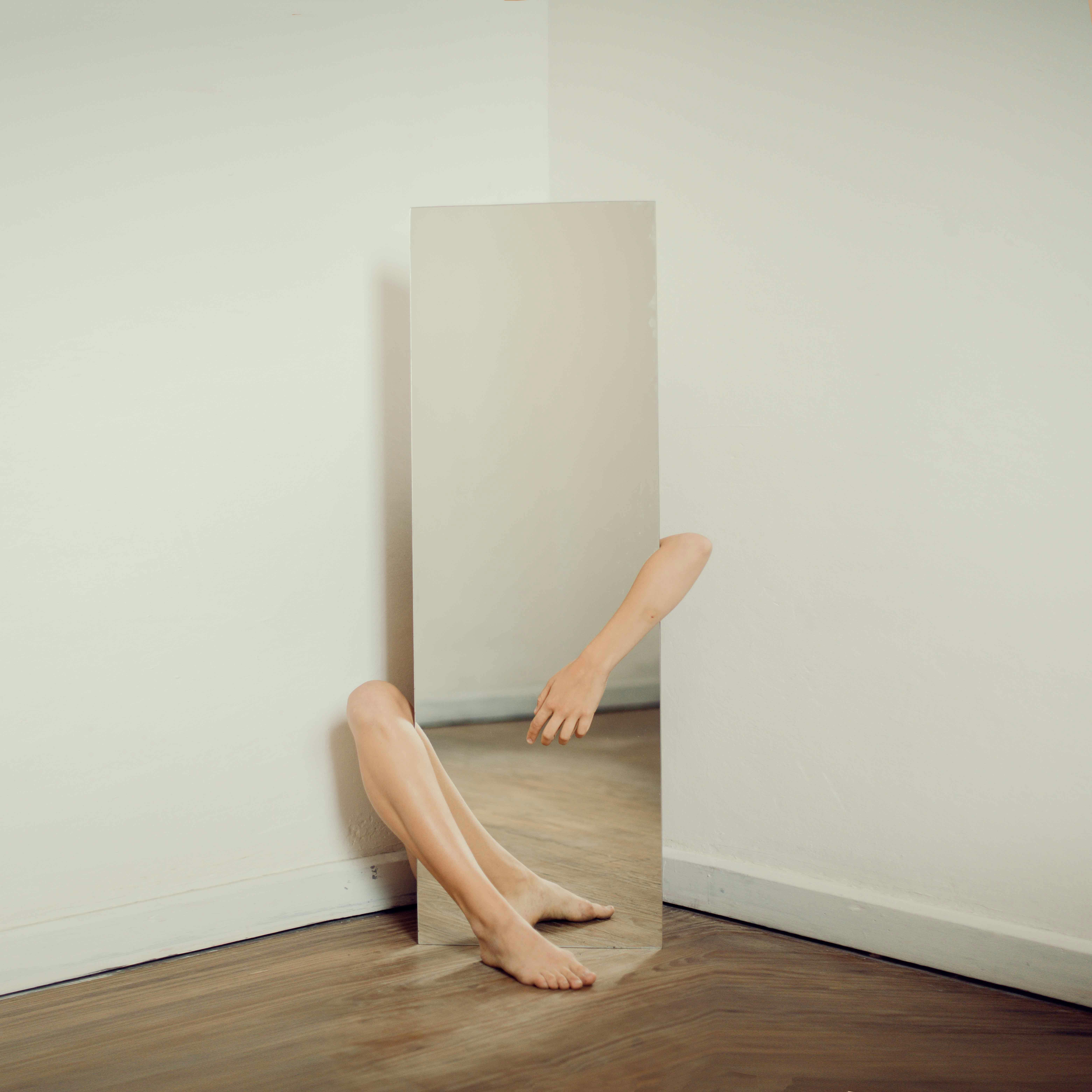 Photograph of a mirror propped in a corner reflecting arms and legs, creating an illusion of a disjointed body